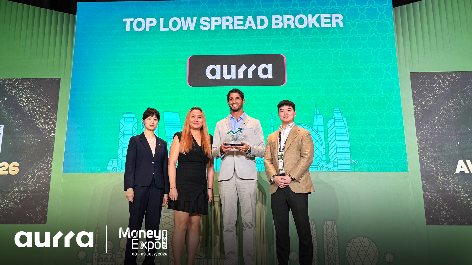 Four representatives stand on stage at the Money Expo Abu Dhabi 2026, holding an award in front of a green screen that reads "TOP LOW SPREAD BROKER" above the "aurra" brand logo.